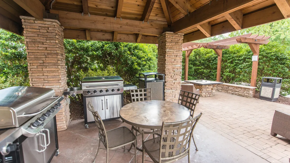 An inviting outdoor BBQ area complete with a modern grill station, a spacious dining table, stone pillars, and a pergola providing shade. The setting is designed for social gatherings, culinary adventures, and relaxed outdoor dining, ideal for evening barbecues or casual get-togethers with friends and family.
