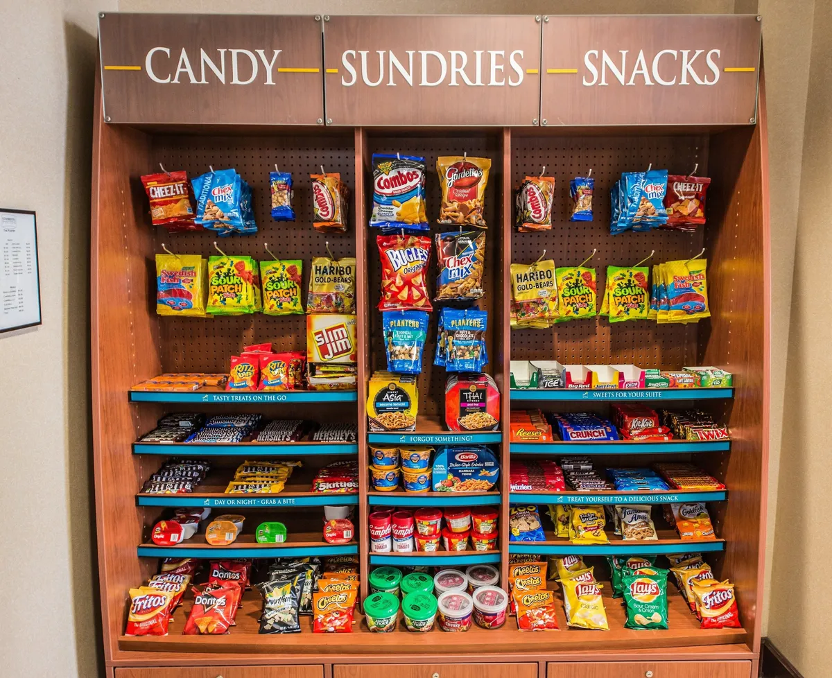 A close-up view of a sundries display filled with colorful candy packages and packaged snack items, set up for easy grab-and-go convenience. The display is appealing and accessible, evoking a sense of comfort and quick refreshment options for residents.