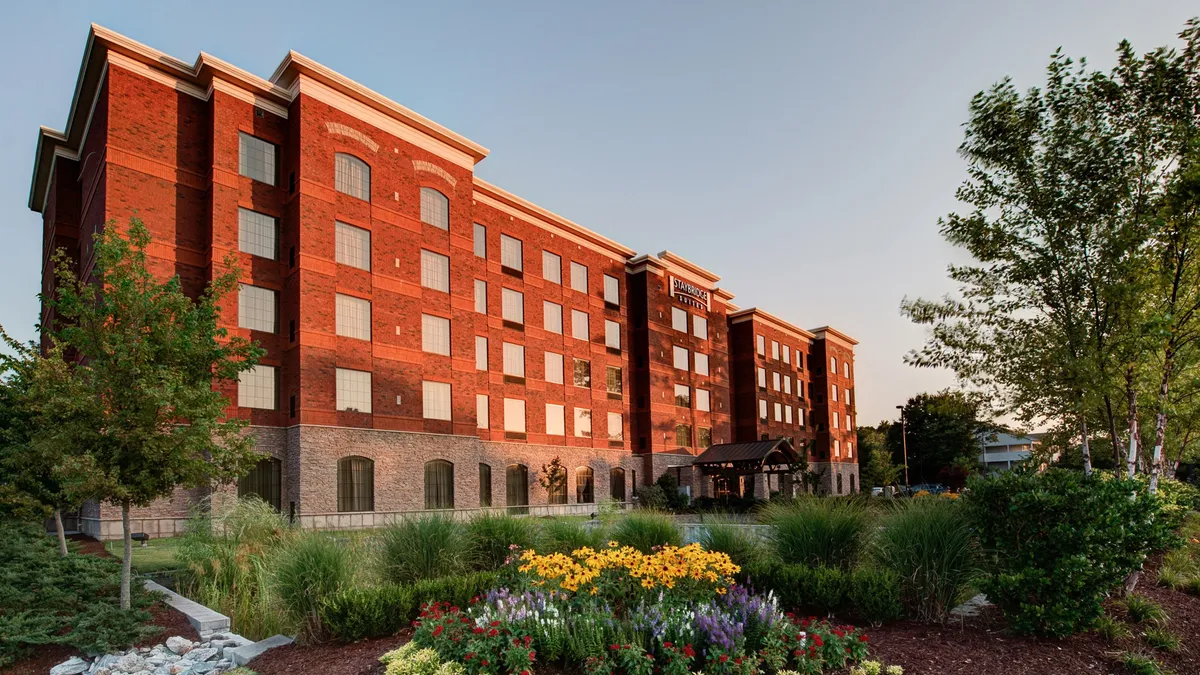 This exterior image shows the welcoming brick facade of the hotel with inviting flower beds and an entrance canopy. Sunlight bathes the scene, highlighting vibrant gardens and the peaceful ambiance of the property’s outdoor space.