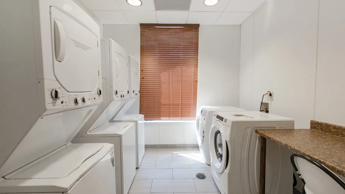 This image features a well-lit laundry room with stacked washer and dryer units, a granite countertop for easy sorting and folding, and a window with blinds that brings in natural light. The setting looks clean, organized, and convenient for extended stays or everyday use.