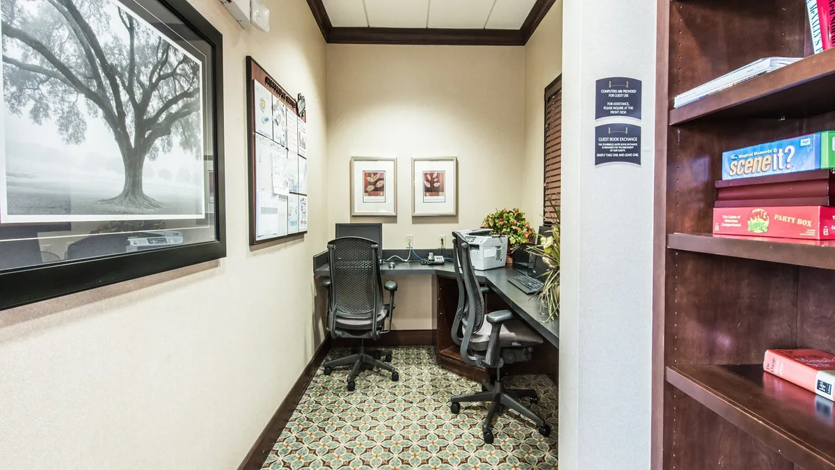 Featuring two desks with comfortable chairs, framed artwork, and shelves with games, this image showcases a productive workspace within a relaxed, inviting environment. The decor is thoughtfully arranged, making the area suitable for focused work or creative pursuits.